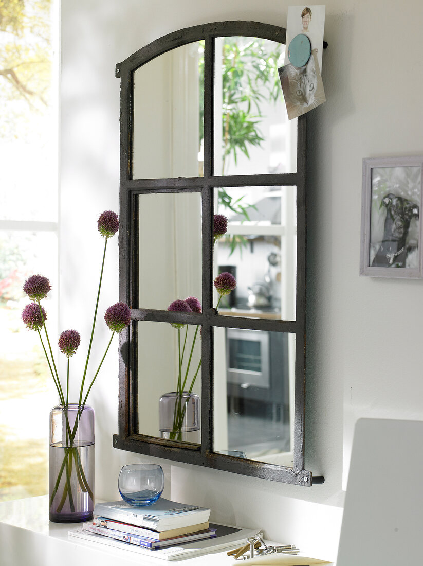 Window mirror on wall with stack of books and flower vase on side table