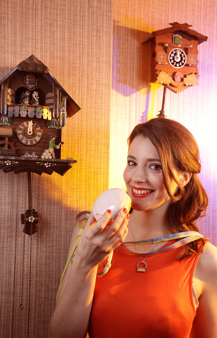 Beautiful woman wearing orange top holding doughnut with cream standing near cuckoo clock