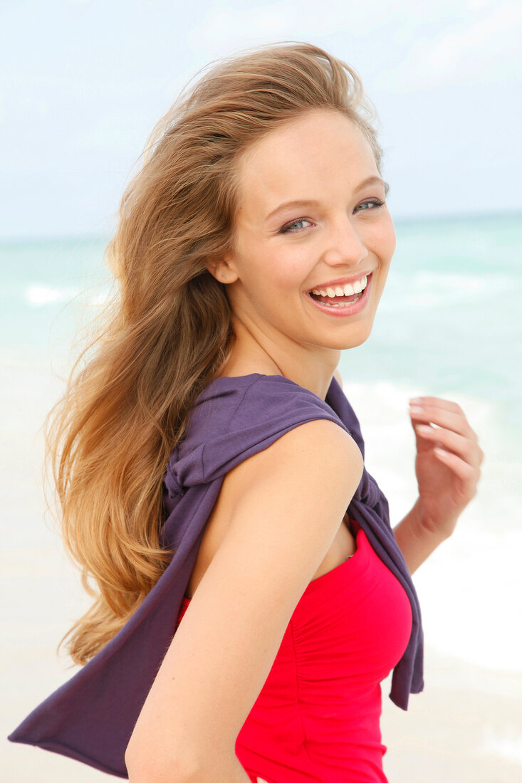 Portrait of beautiful blonde woman wearing red dress standing on beach, smiling