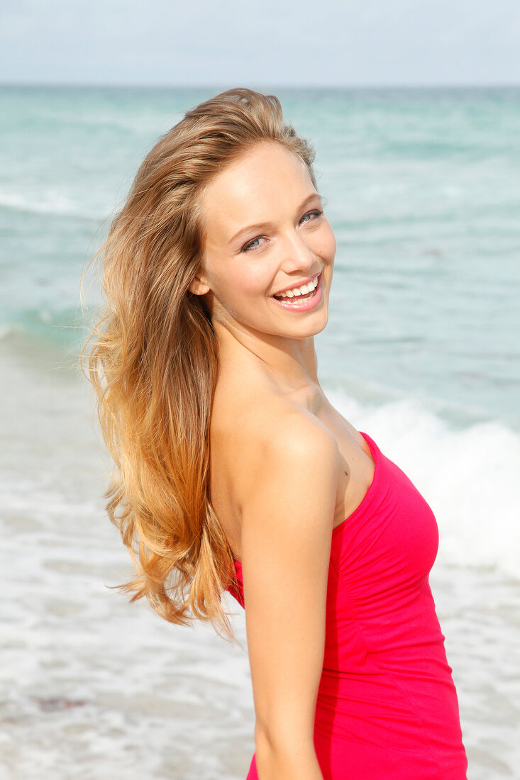 Portrait of beautiful blonde woman wearing red dress standing on beach, smiling