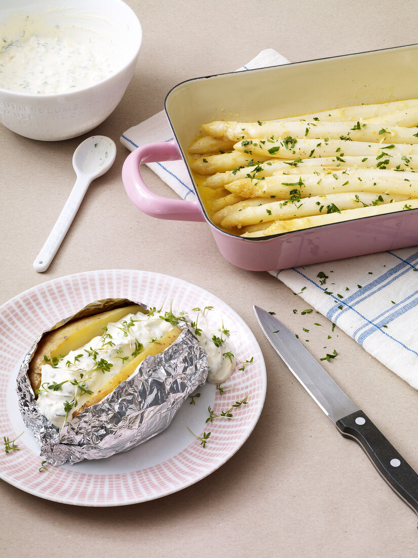 Oven asparagus in baking dish and baked potato with green sauce on plate