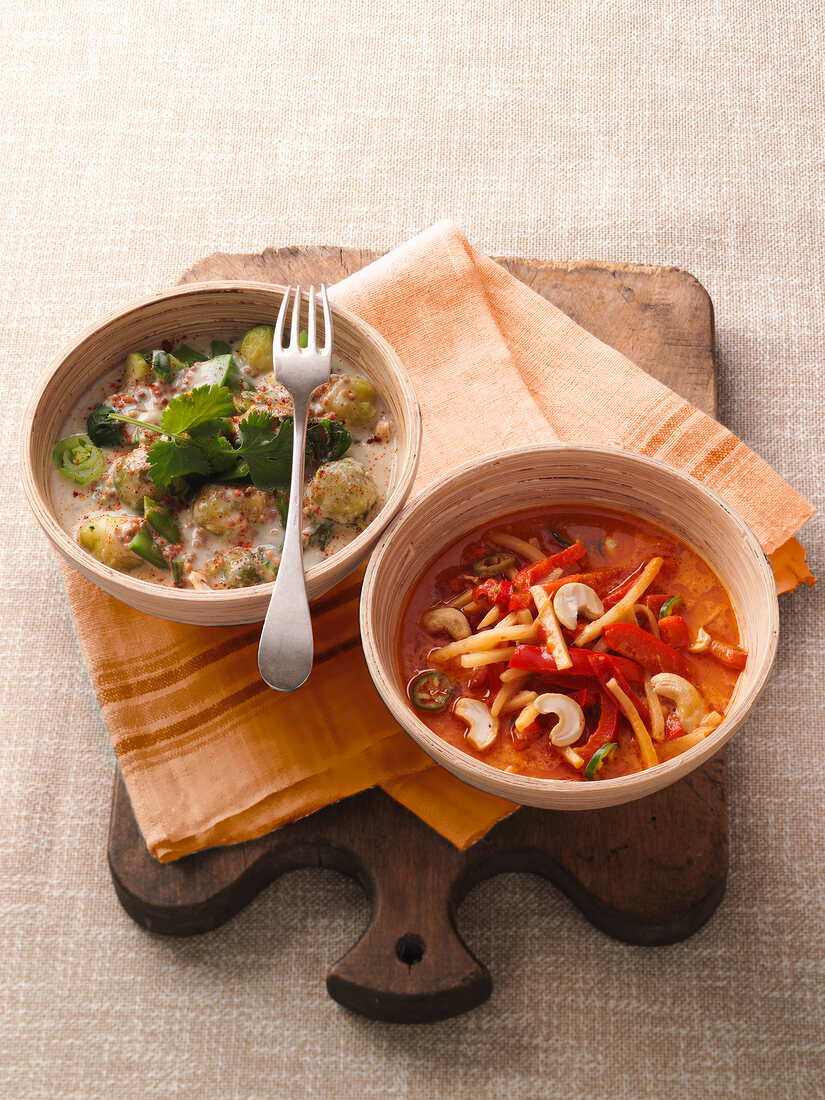 Two bowls of green vegetable curry and red curry on wooden serving board