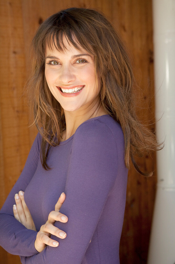 Portrait of happy brown haired woman in purple top standing against wooden wall, smiling