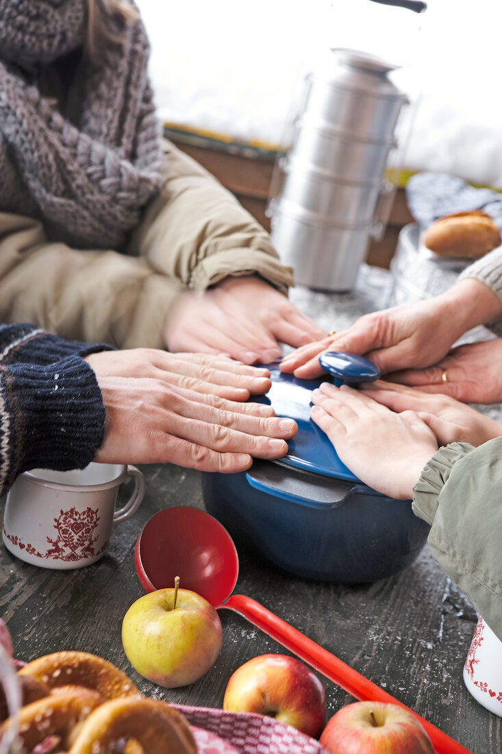 People warming hands on bowl of hot soup with lid on table with apples and ladle