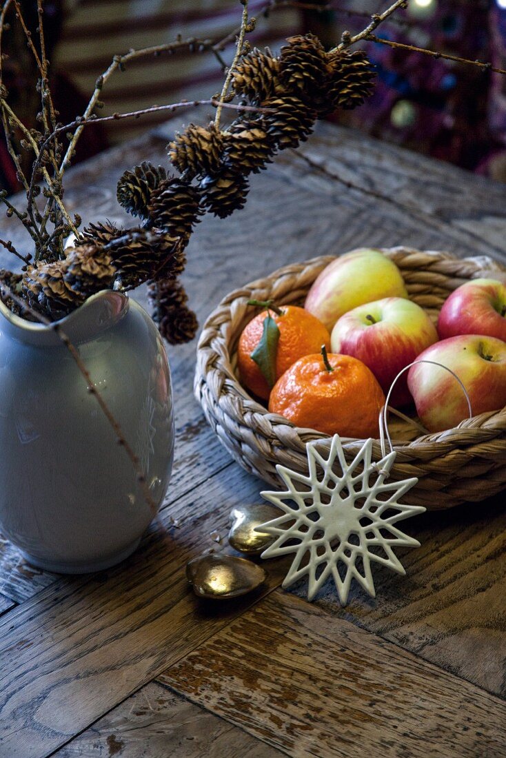 A basket of fruit next to a vase with larch twigs