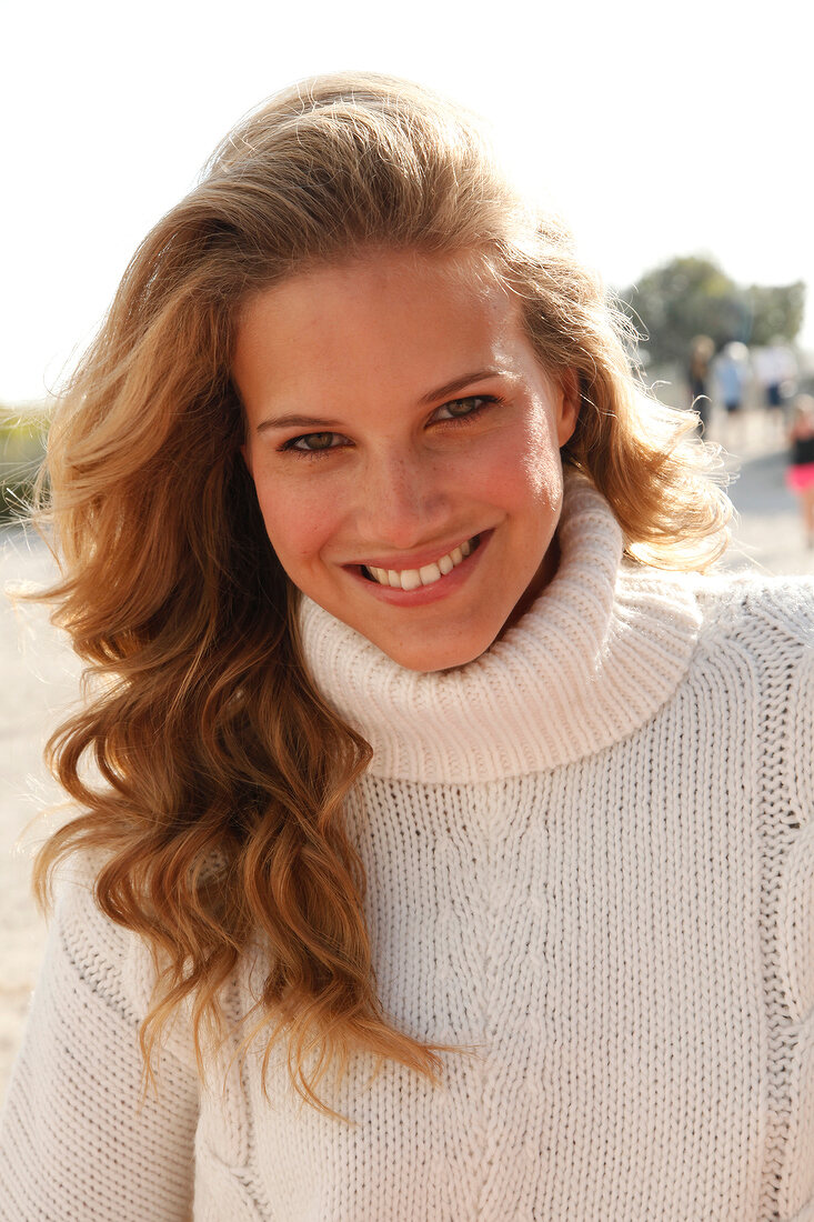 blonde Frau in weißem Pulli am Strand, lächelt in Kamera