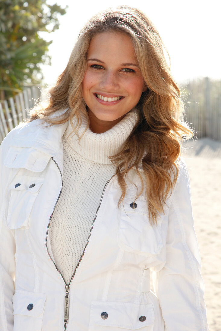 Pretty blonde woman wearing white jacket over white sweater standing on beach, smiling