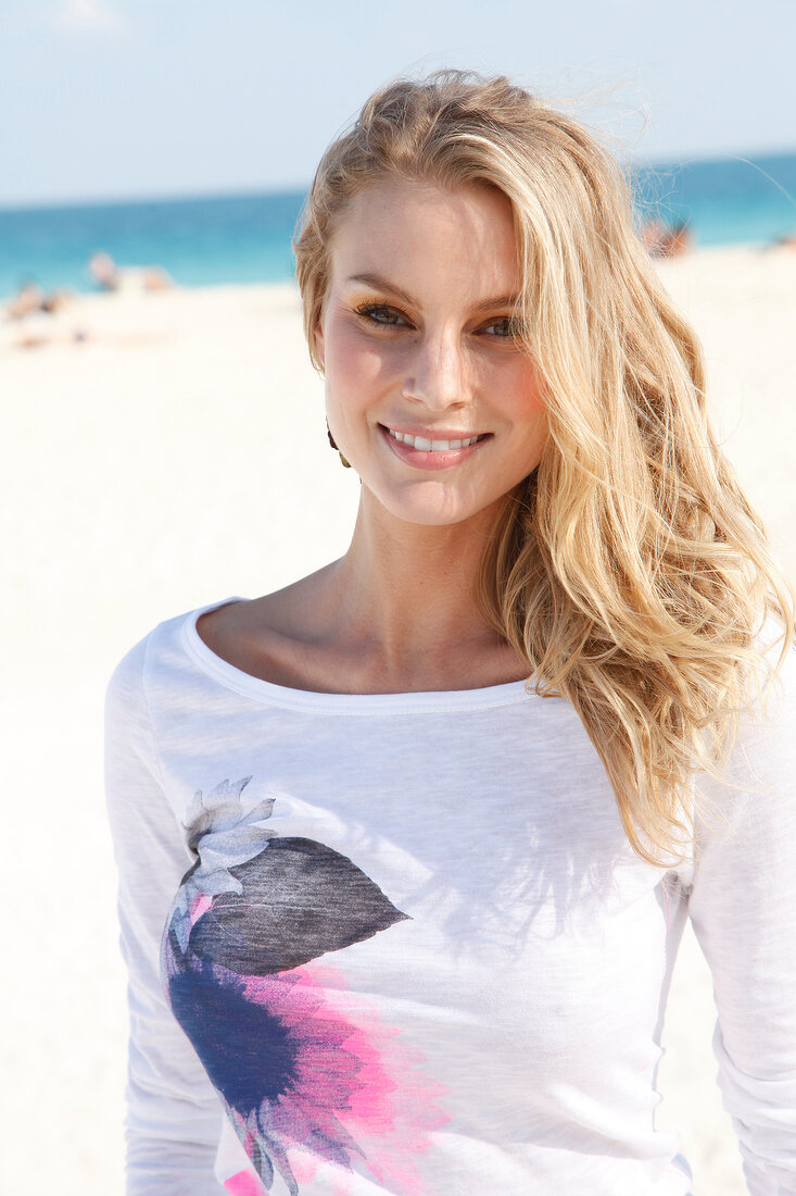 Portrait of beautiful blonde woman wearing white top standing on beach, smiling