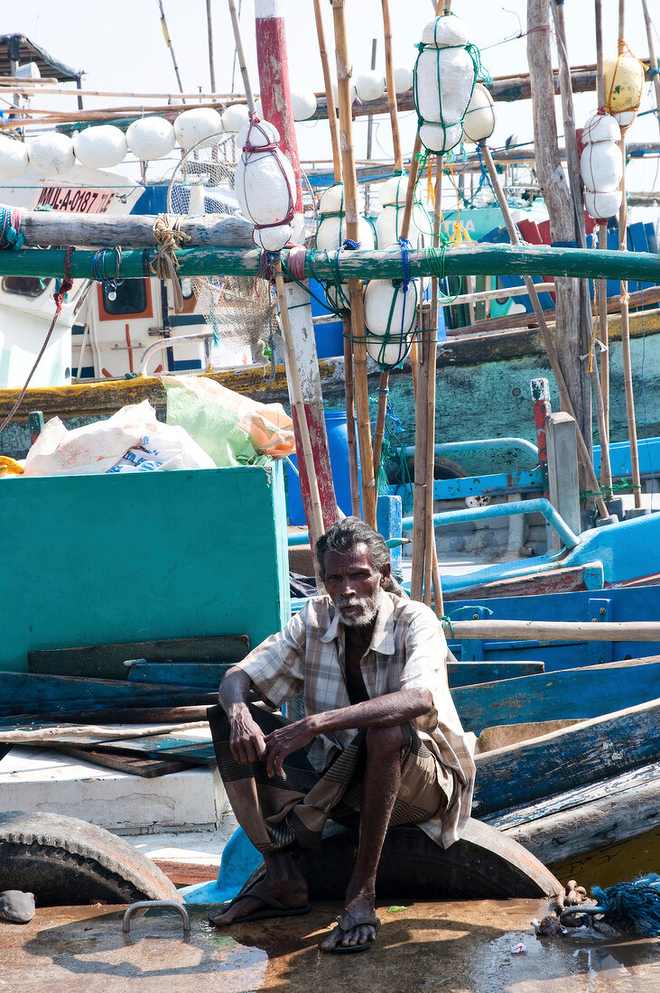 Senior man sitting on harbour with fishing boats in background in Tangalle, Sri Lanka
