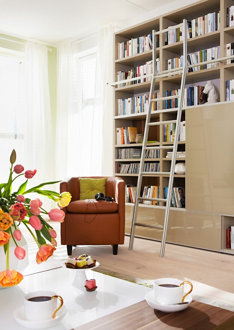 Ledersessel vor Bücherregal mit Leiter im Wohnzimmer, im Vordergrund Couchtisch mit Tulpen und Kaffeetassen