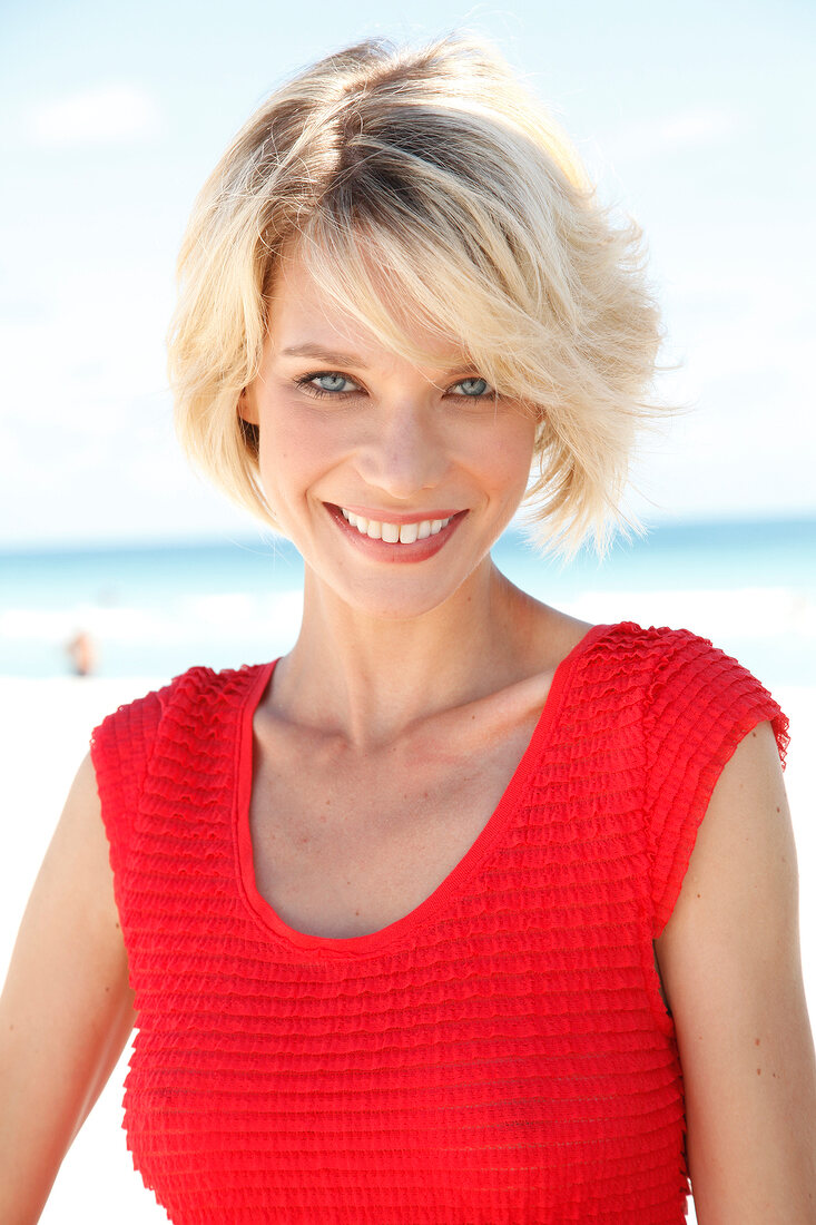 Portrait of pretty blonde woman with short hair wearing red top sitting on beach, smiling