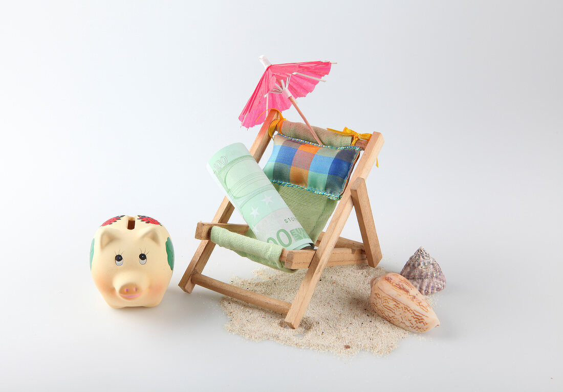 Rolled bill on deck chair with umbrella, mussels , sand and piggy bank on white background