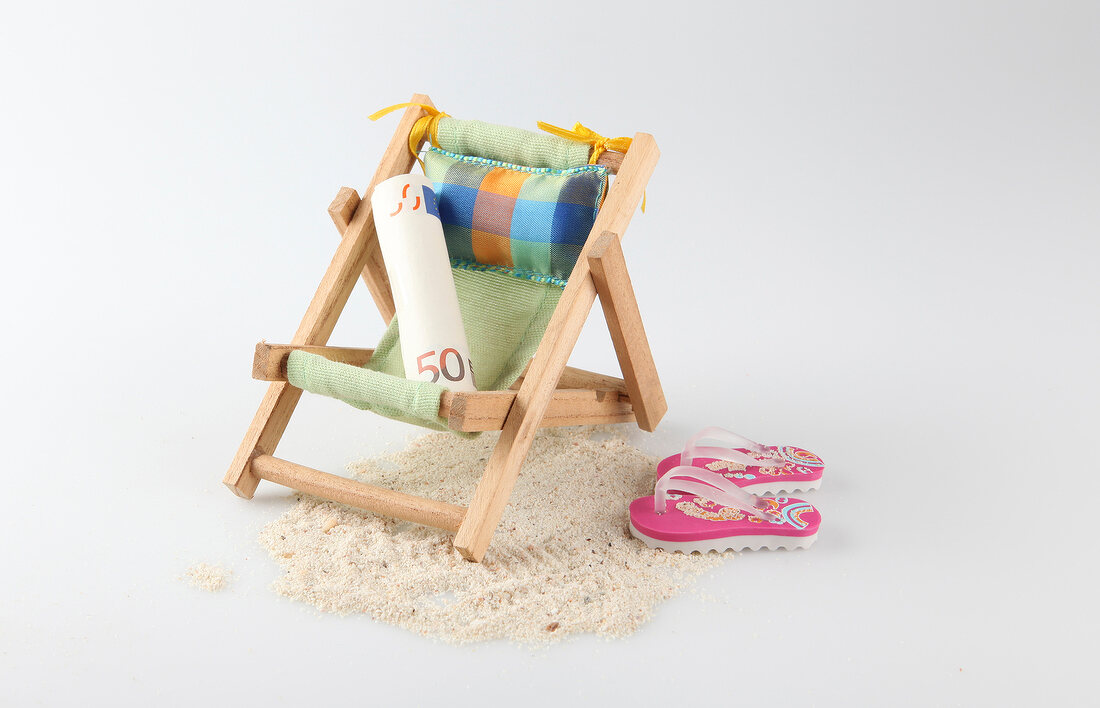 Rolled-up currency on small deck chair with flip-flops and sand