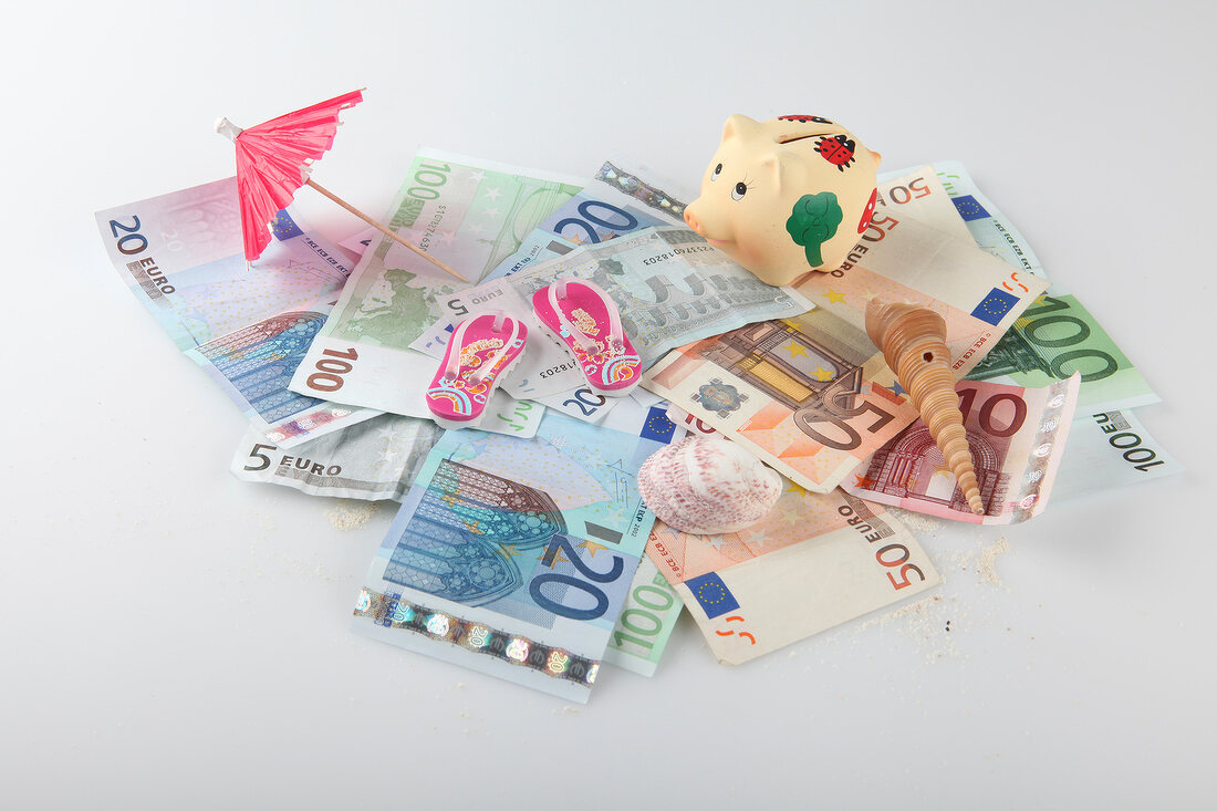 Heap of bills, umbrella, flip-flops, shells and piggy bank on white background