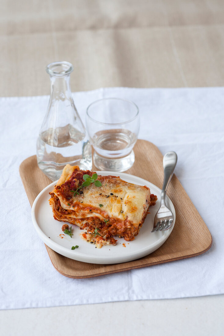 Lasagne al forno on plate with flask and glass of water on wooden serving tray