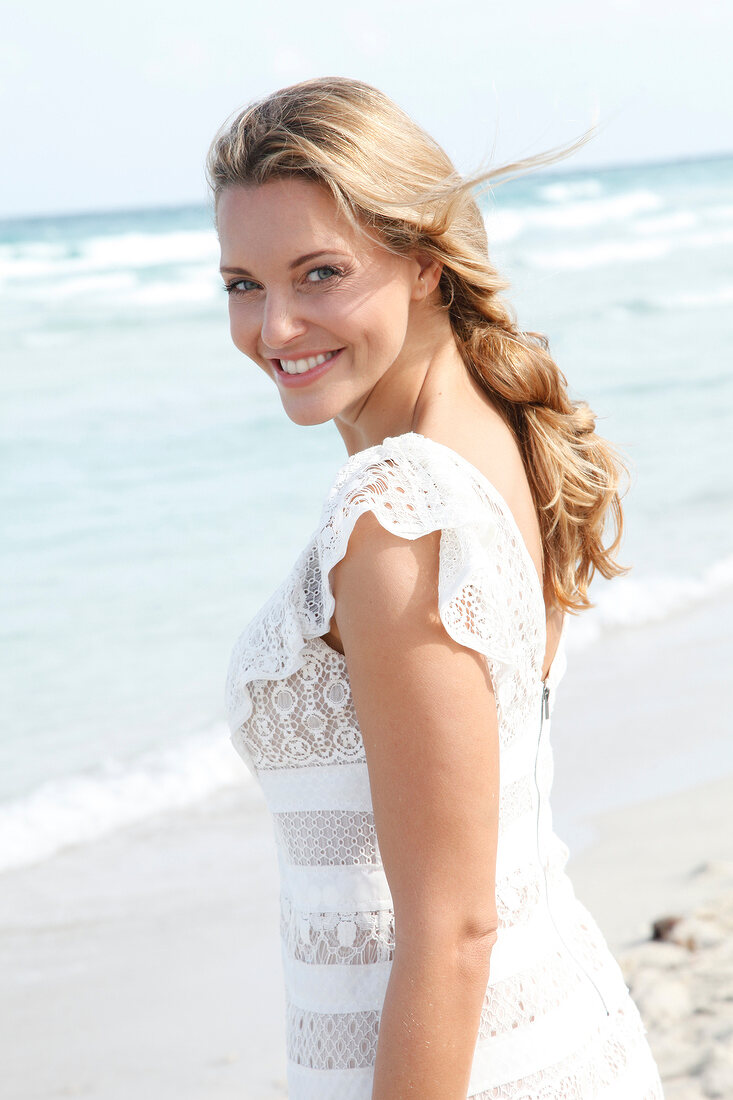 blonde Frau im weißen Kleid am Strand, lacht in die Kamera