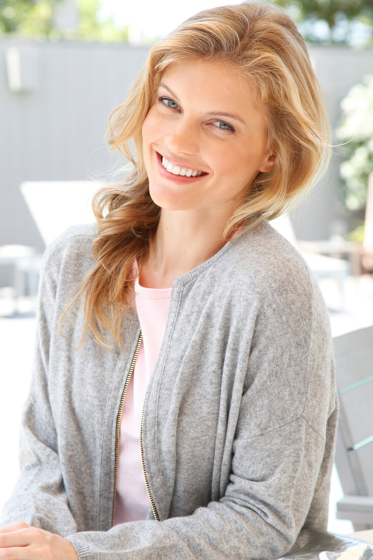 Portrait of blonde woman wearing pink top and gray jacket standing at poolside, smiling