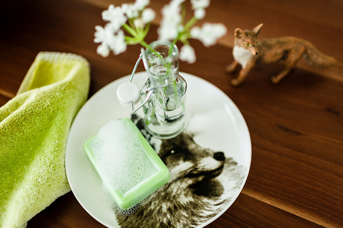 Green soap and flower in glass bottle in plate with green towel on wooden surface