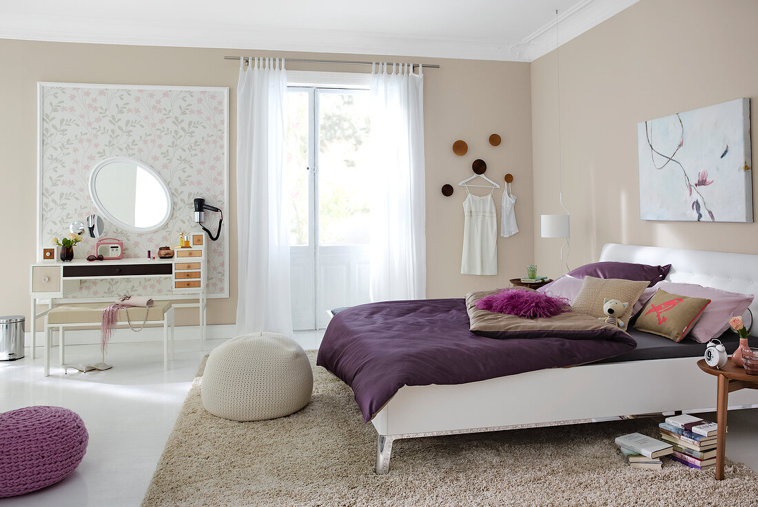 Bedroom with beige walls, bed with purple bed blanket, cushions and dressing table