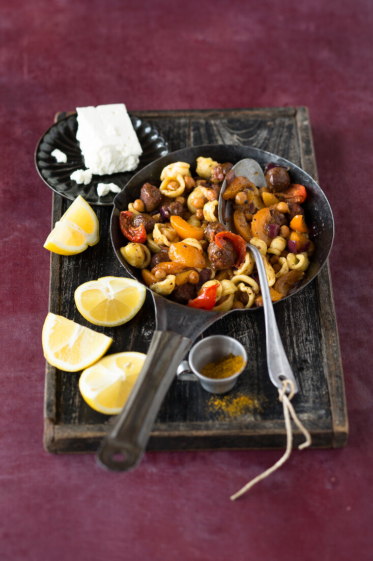 Orecchiette and merguez in pan beside lemon wedges on wooden tray