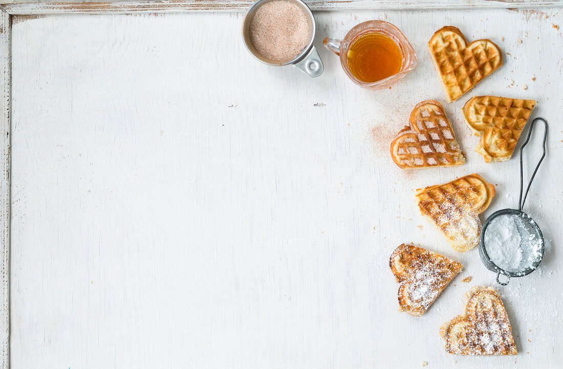 Heart shaped waffles with icing sugar in sieve and maple syrup in jug on white background