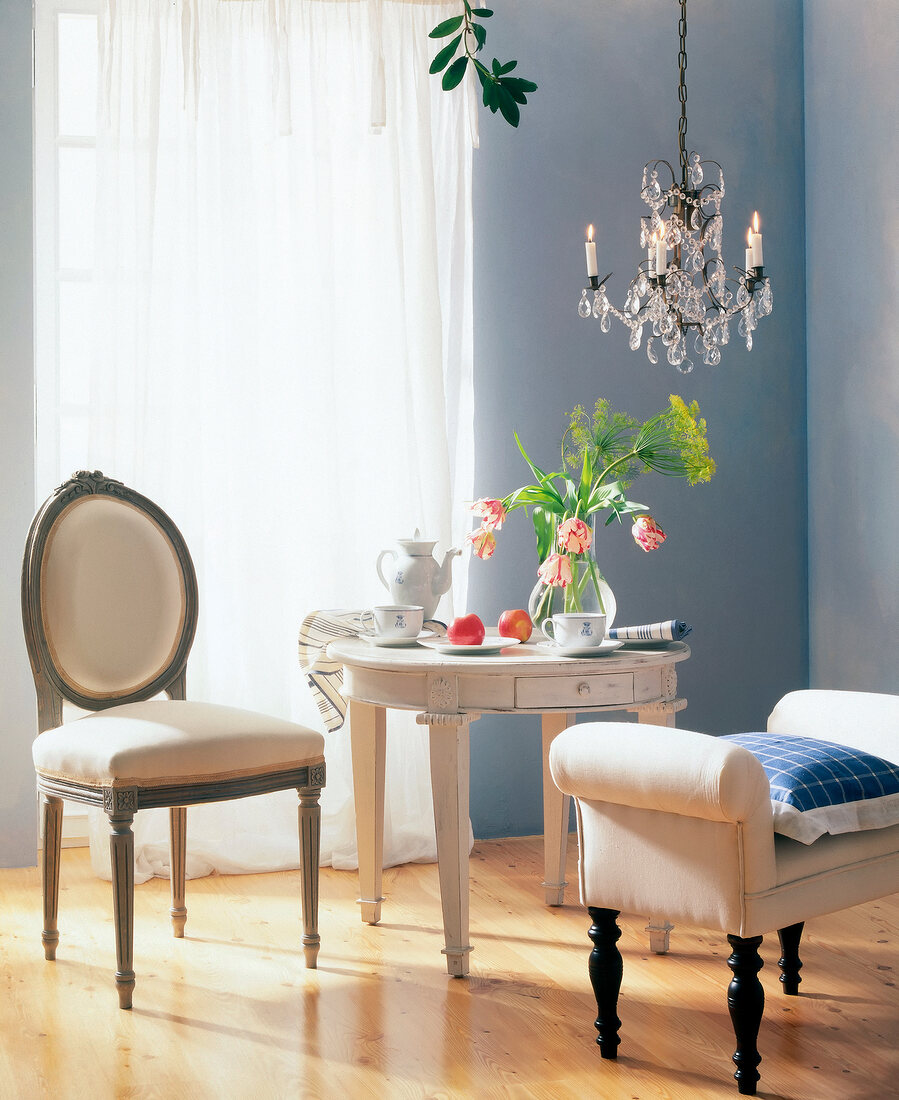Room with crystal chandelier, wooden floor, table, stool and chair in pastel shades