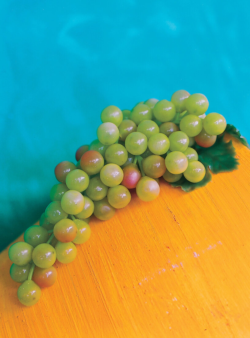 grüne künstliche Weintrauben liegen auf gelbem Tisch vor blauer Wand