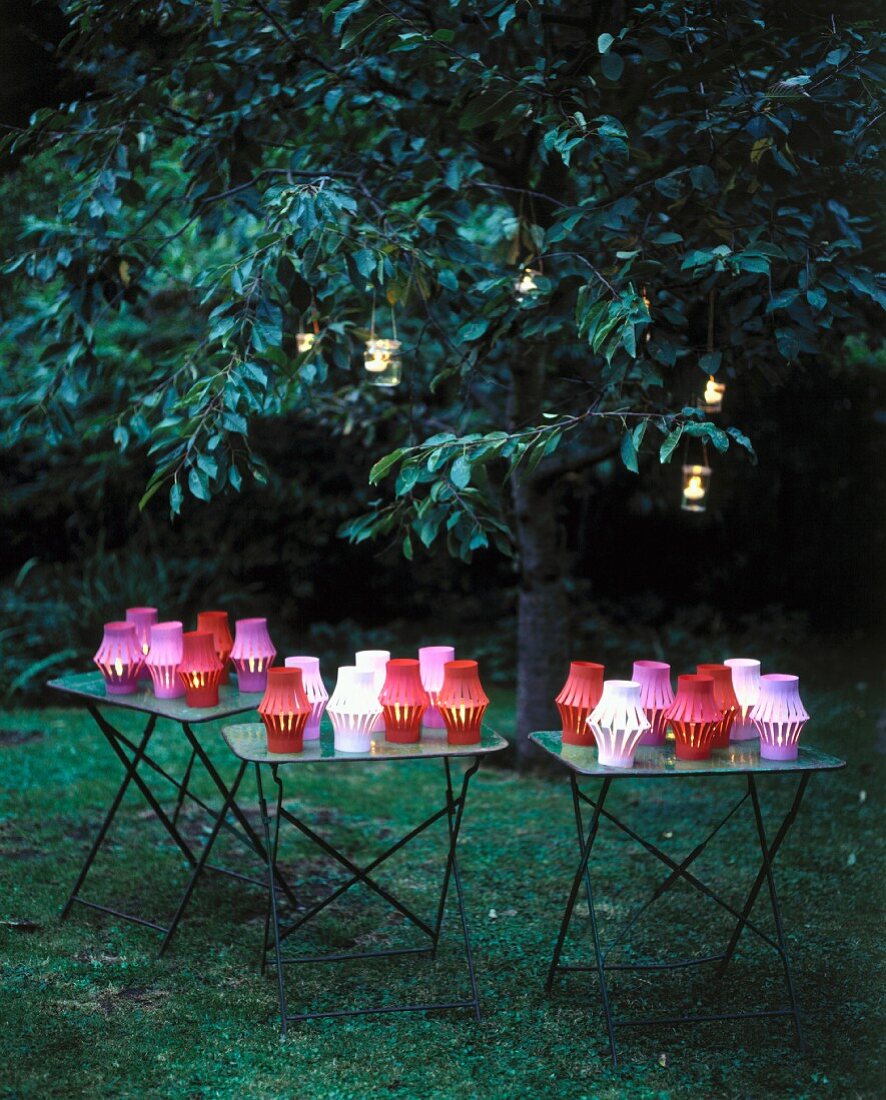 Verschiedenfarbige leuchtende Papierlaternen auf Tischen und in Bäumen, im Garten mit Abenddämmerung