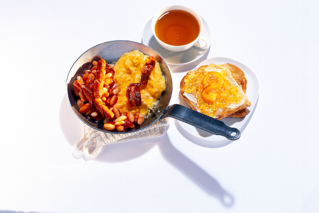 Breakfast table set with scrambled eggs, beans, bacon, tea, toast with jam on table on mat