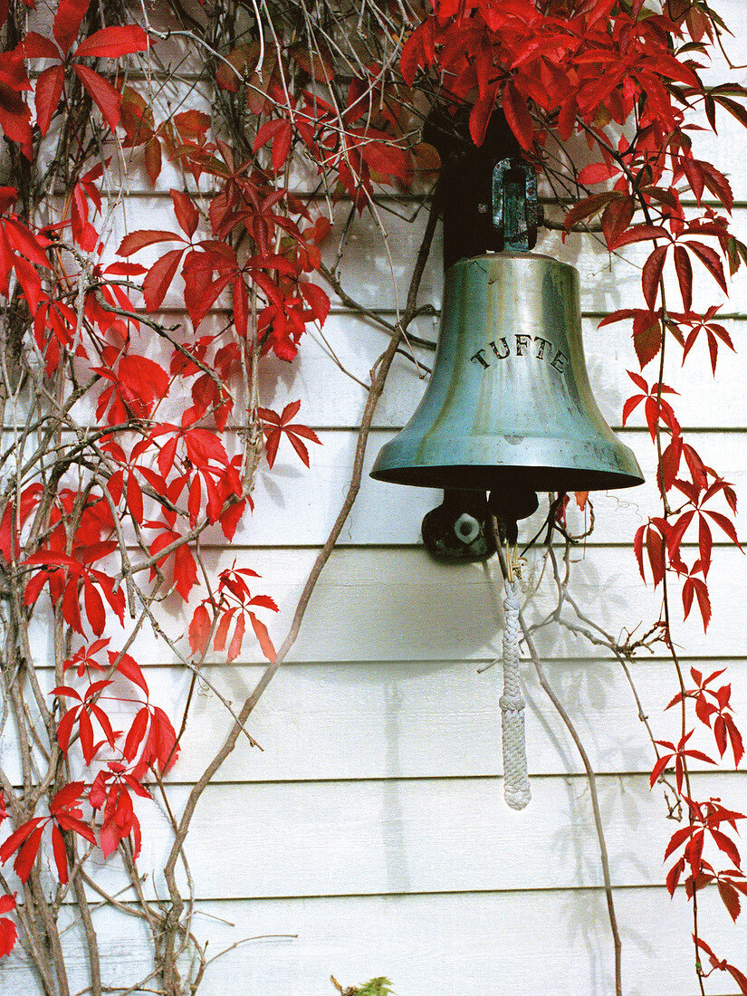 Bell hanging in front of wooden house covered with red vine leaves