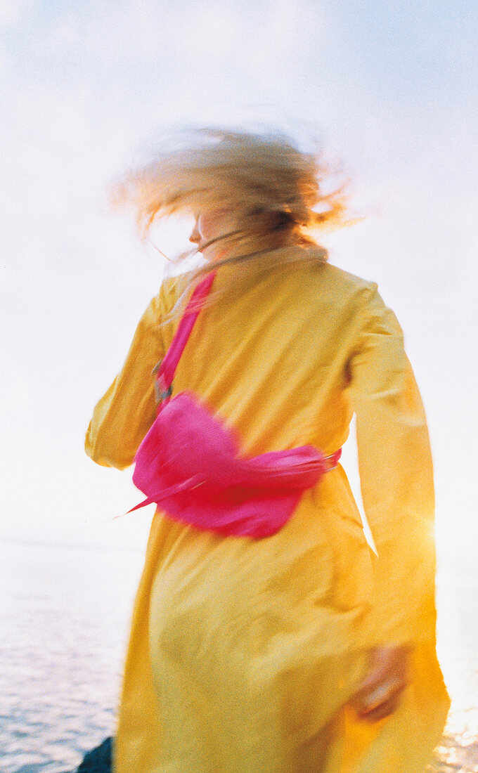 Frau im gelben Mantel + Tasche in pink am Meer, Bewegungsunschärfe