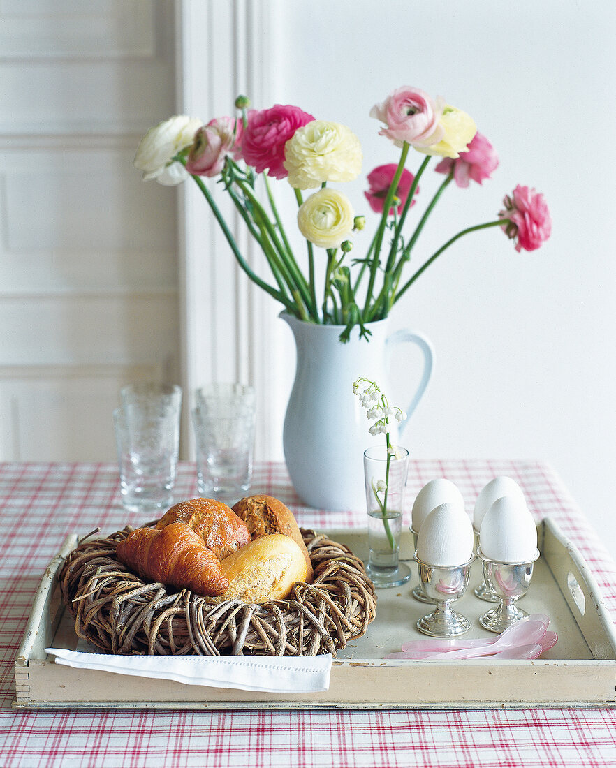 Breakfast with bread and croissants in basket and eggs on tray, ranunculus in vase