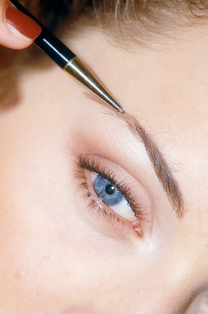 Close-up of blue eyed woman darkening her eyebrow with eyebrow pencil