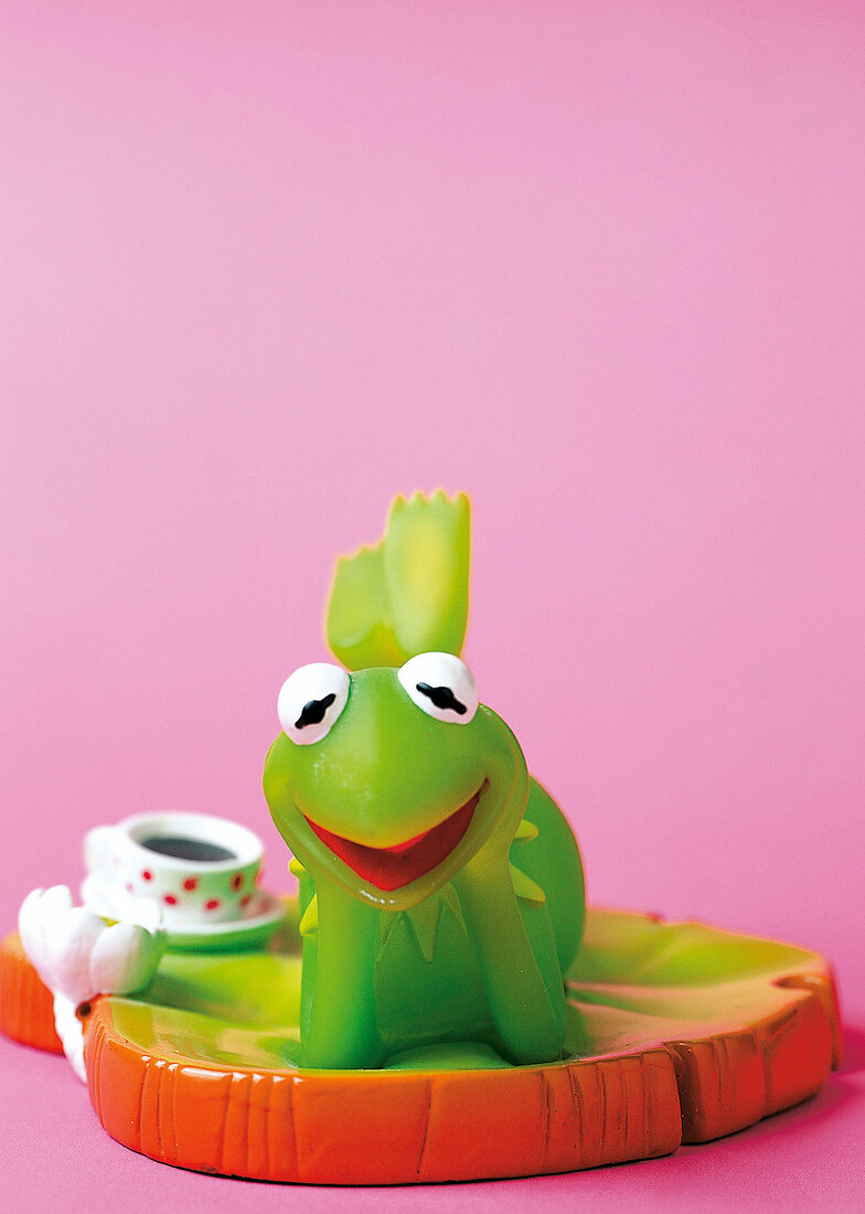 Kermit der Frosch liegt auf einem Blatt mit einer Tasse im Hintergrund