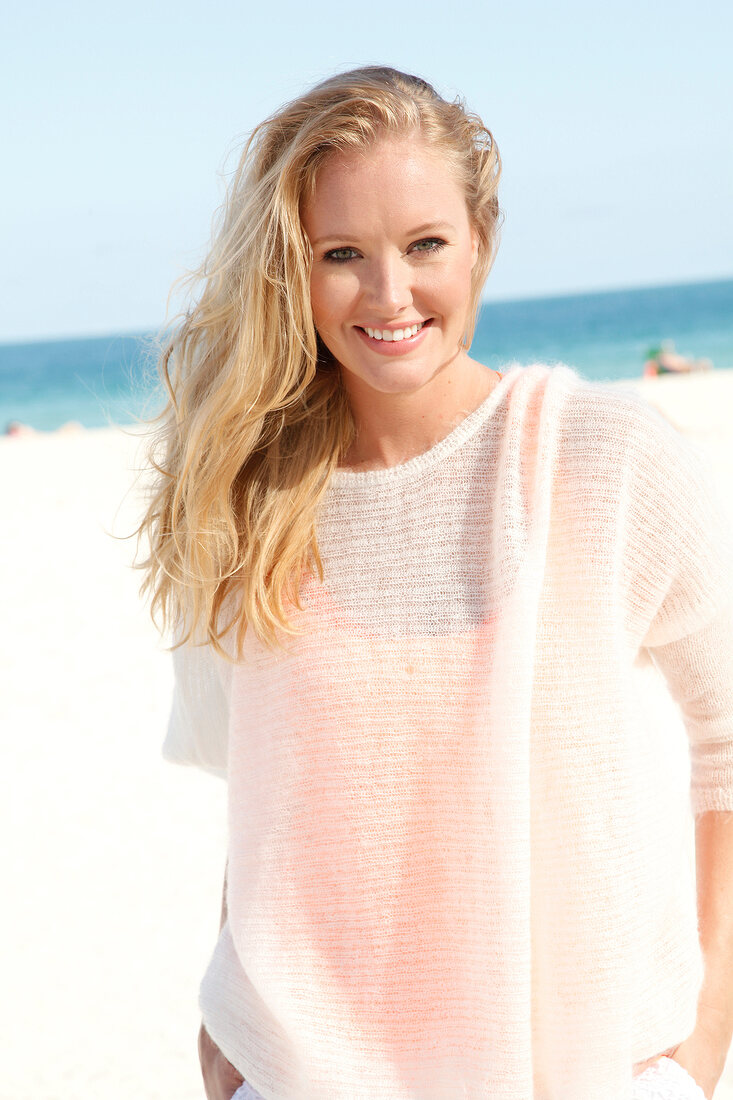 Blonde woman in a white sweater on the beach, smiling at camera