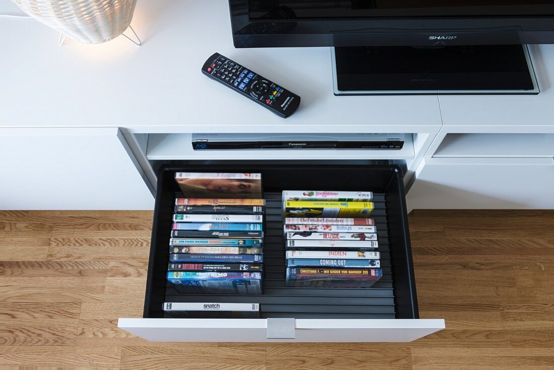 A collection of DVDs in an open drawer in a media unit with a television and a remote control
