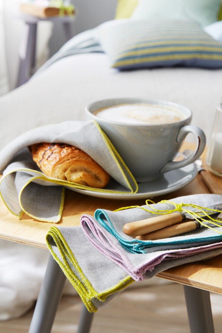 Pain au Chocolat in Stoffserviette neben Cappuccino und Serviettenbündel auf Holzstuhl vor einem Bett