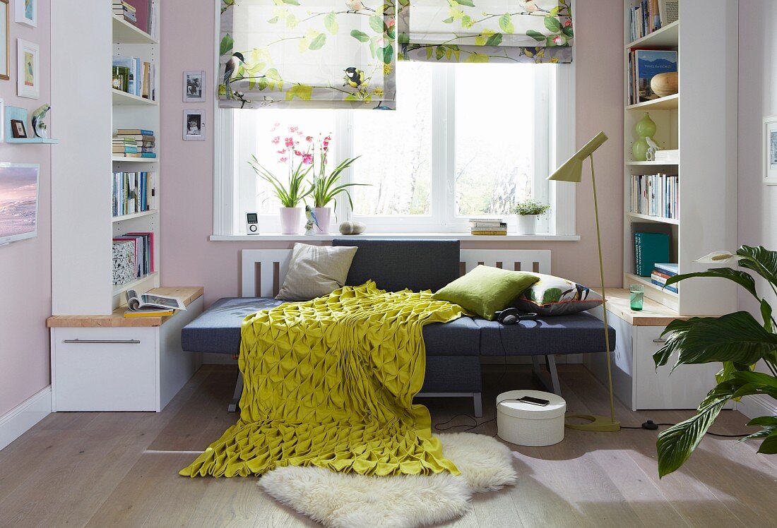 An armchair folded out into a bed with cushions and a green-and-yellow structured blanket between bookshelves