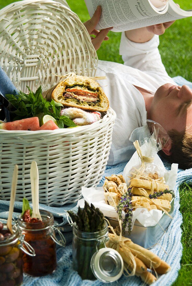 A full picnic basket next to a man lying on the picnic blanket reading a book