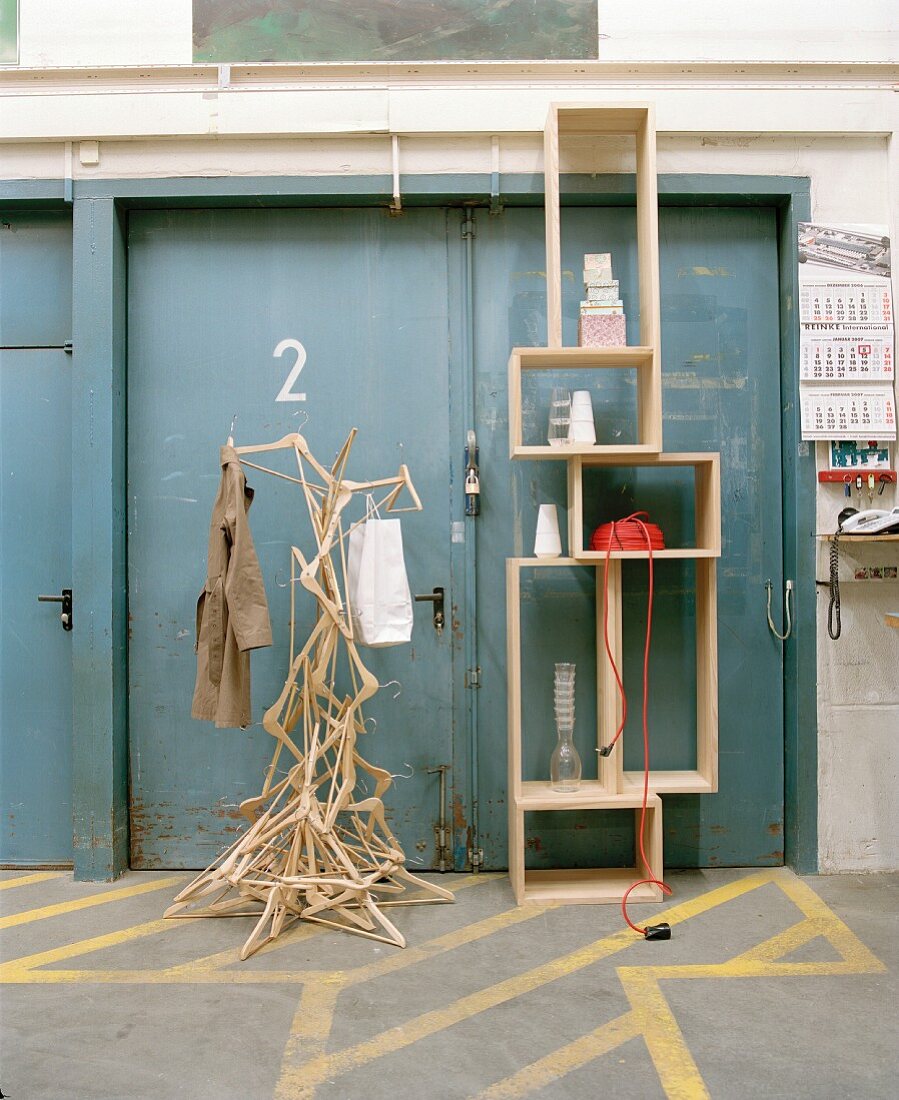 Künstlerisch gestaltete Garderobe aus Kleiderbügeln neben modernem Regal aus Holz vor grauen Metalltüren