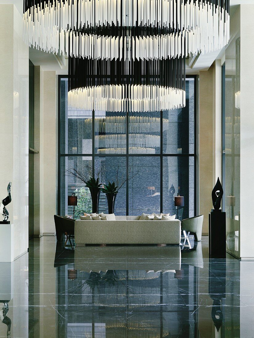 Reception area with circular hanging lamps made of illuminated rods and white upholstered sofa in front of a bank of windows on a reflective stone floor