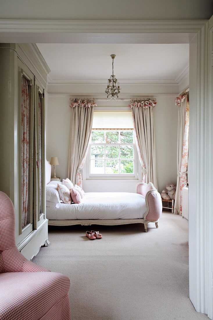 Feminine bedroom in soft shades of pink with old, white wardrobe, double bed with upholstered footer and pretty, gathered pale curtains in background. Elegant country-house ambiance.