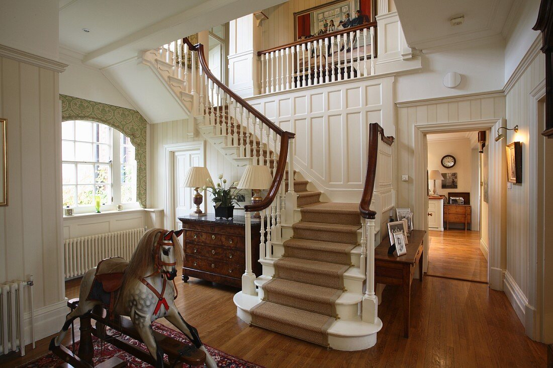 An antique rocking horse in front of a white-painted wooden staircase in the spacious hallway of a classy but comfortable country house with parquet flooring