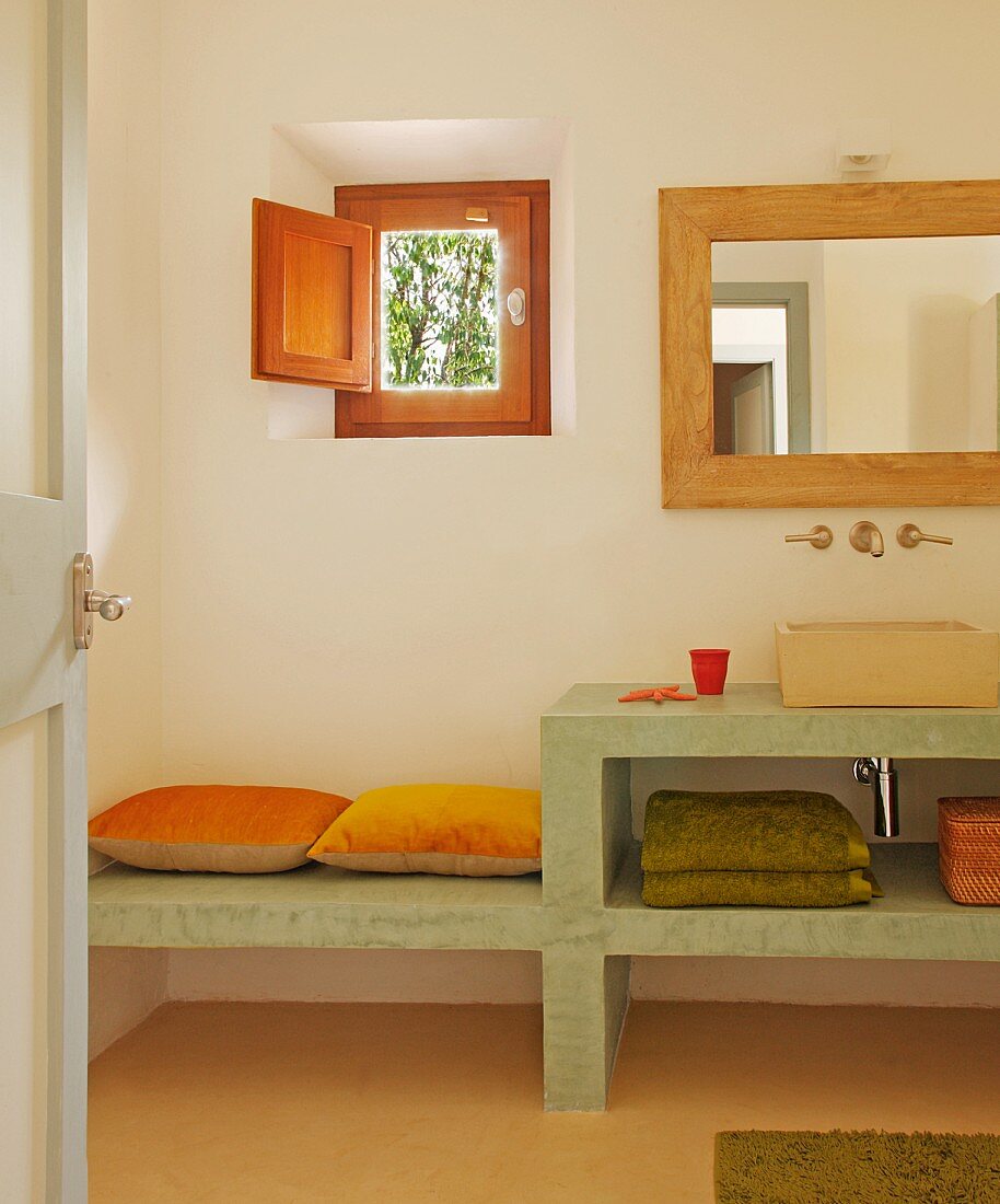 Blick durch offene Tür auf schlichten Waschtisch aus Beton mit integrierter Bank an Wand unter kleinem Fenster mit Holzladen