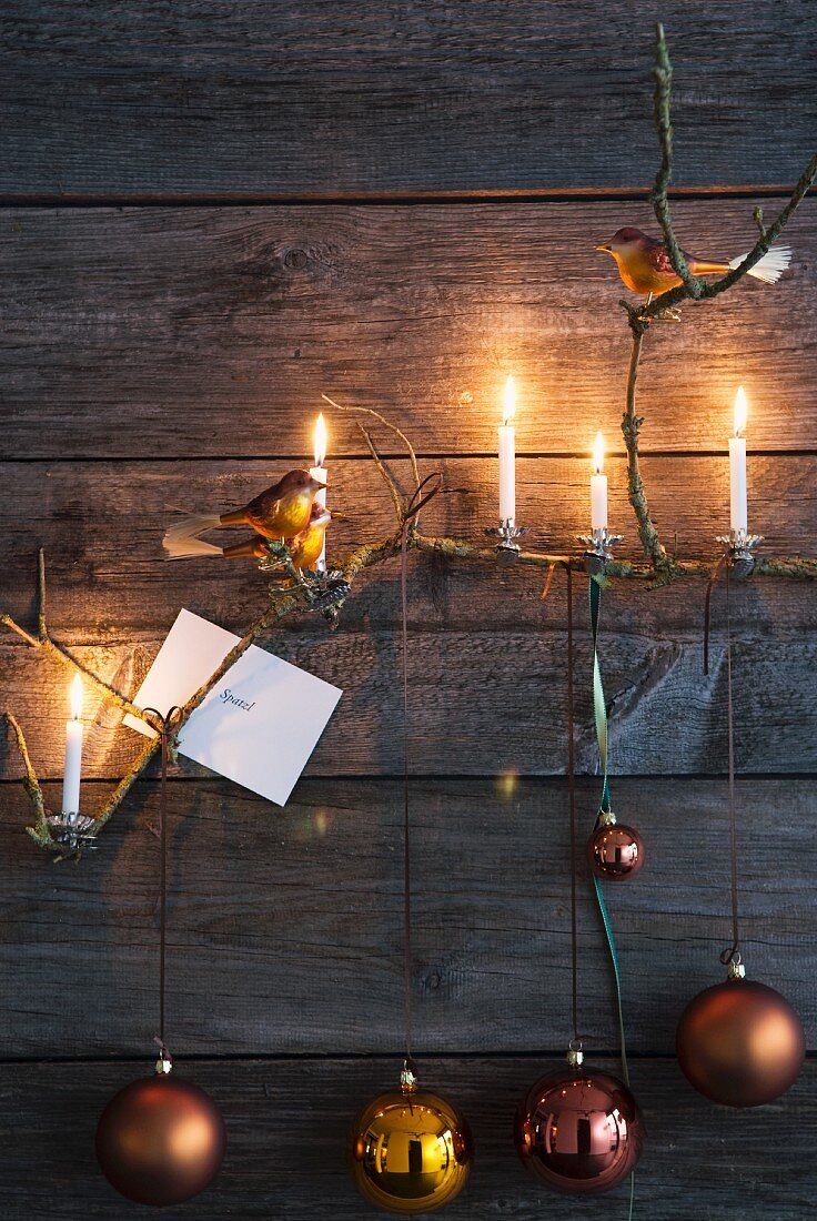 An Holzwand aufgehängte Weihnachtskugeln und brennende Kerzen an Zweig im Kerzenhalter