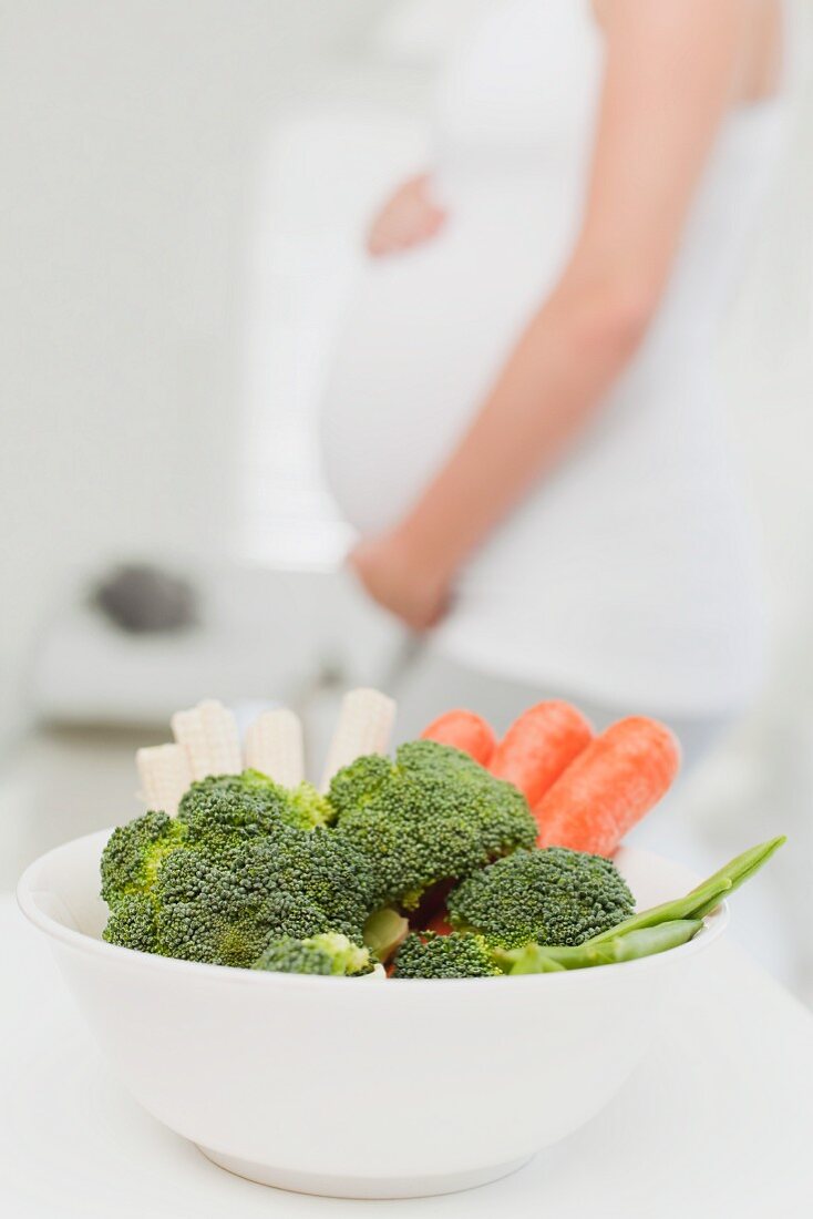 Eine Schüssel mit frischem Gemüse, dahinter eine schwangere Frau