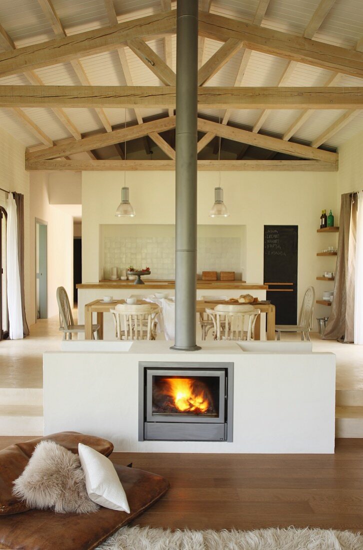 Masonry fireplace built in centre of open-plan interior in front of dining area and kitchen on platform in Mediterranean house