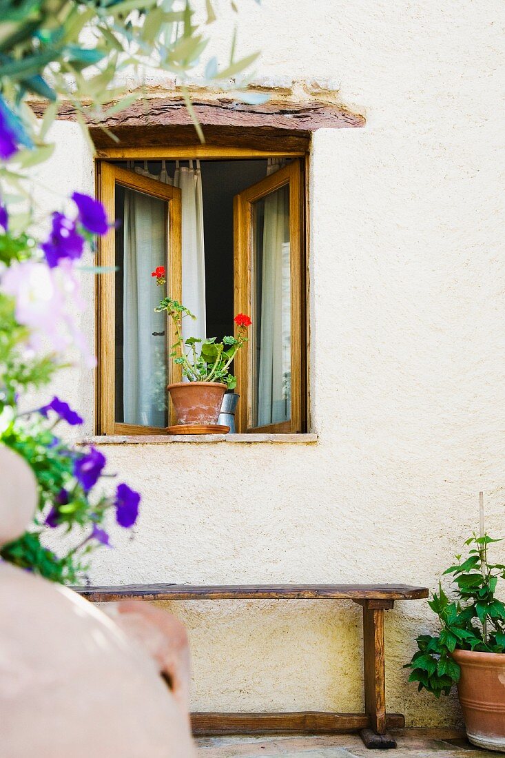 Eine Holzbank an der Hauswand unter dem geöffneten Fenster mit Geranie