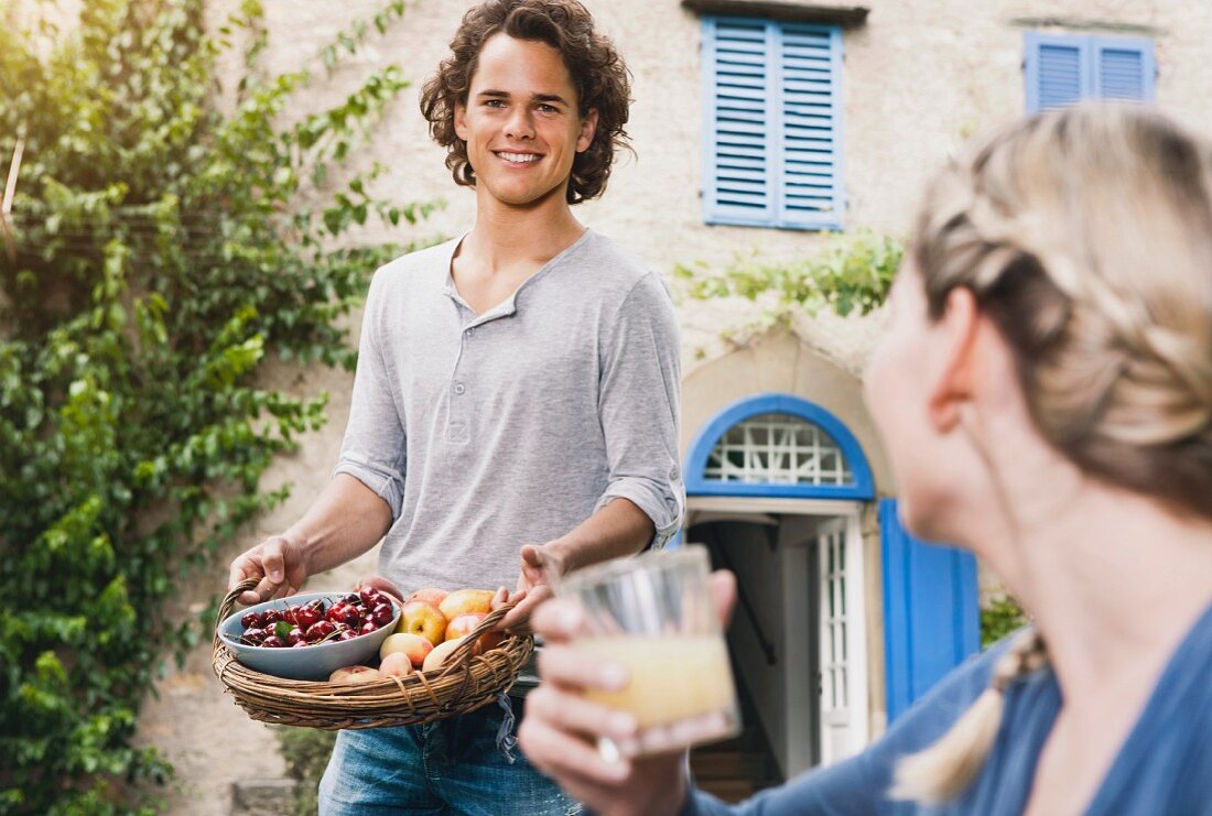 A young man carrying a basket of apples and cherries and a young woman drinking a glass of apple juice