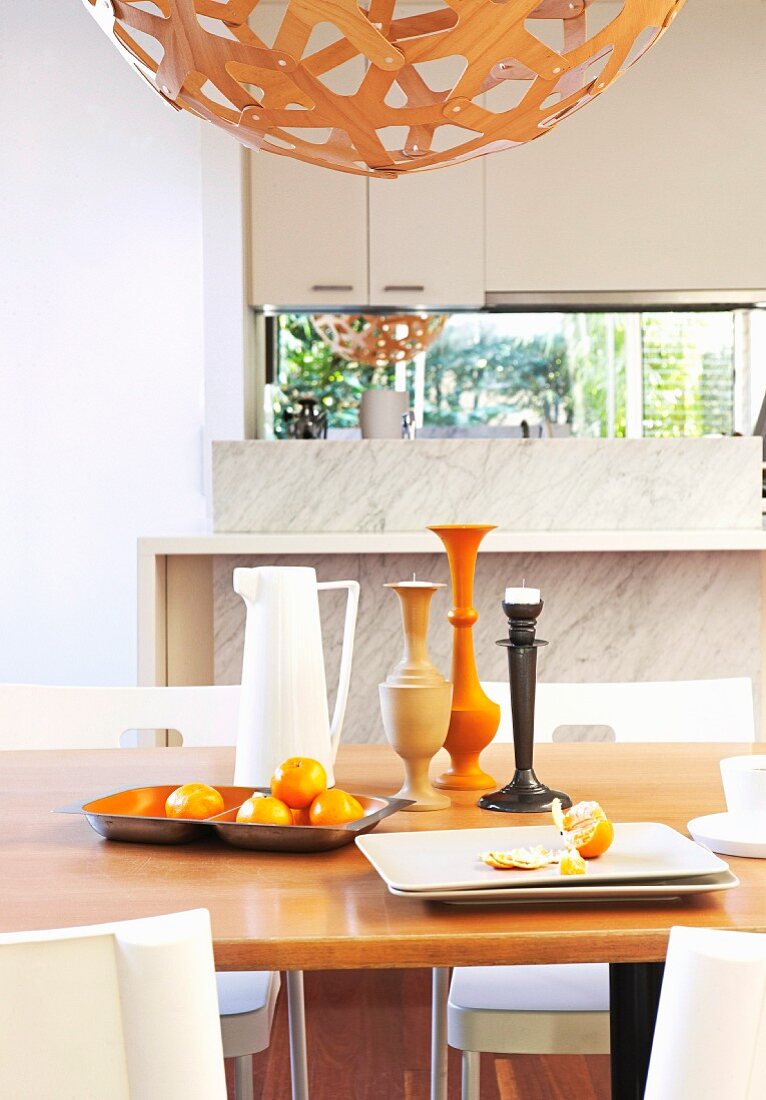 Still-life: fruit bowl, jug and candlesticks on wooden table below partially visible, spherical pendant lamp in dining area